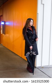 Vertical View Of Pretty Brunette Woman With Waist Length Hair Wearing Black Coat Standing With Dejected Expression Holding An Umbrella At The Entrance Of An Indoor Parking Building