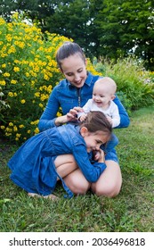 Vertical View Of Pretty Brunette Young Woman Kneeling In Grass Holding Happy Baby, With Older Sister Grabbing Her Knees, Quebec City, Quebec, Canada