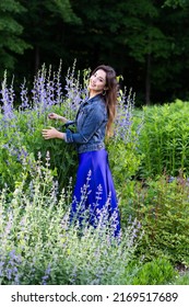 Vertical View Of Pretty Smiling Brunette Woman In Long Blue Dress And Denim Jacket Standing In Garden Among Russian Sage In Bloom