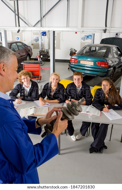 A vertical view of a teacher standing\
in the foreground holding an auto part while teaching automotive\
trade to his students in the vocational\
school