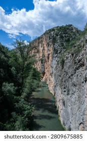 Vertical view of the Turia river flowing through a deep limestone canyon with massive steep cliffs under a cloudy blue sky in Chulilla near Valencia, Spain.
