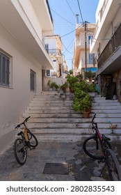 Vertical view of two bicycles on a paved street at the base of historic whitewashed stairs decorated with potted green plants and blooming flowers, leading up through old houses in Crete.
