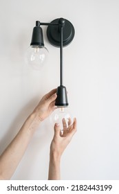 Vertical view of woman replace broken light bulb in black metal sconce. Electrician change energy saving lamp in apartment with white living room wall. Energy efficient equipment, household fixture