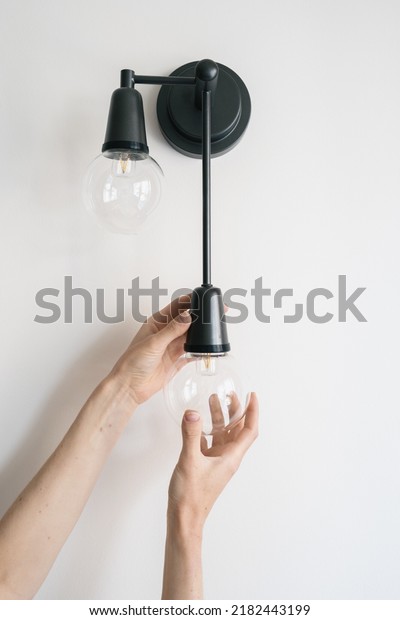 Vertical view of woman replace broken light bulb\
in black metal sconce. Electrician change energy saving lamp in\
apartment with white living room wall. Energy efficient equipment,\
household fixture