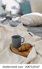 Vertical view of wooden breakfast tray with coffee cup and yummy baked croissant in cozy bedroom. Concept of lazy weekend morning at home with delicious food in bed