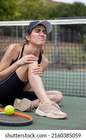 Vertical View Of Young Woman In Pain With Injured Knee. With Pain Face Almost Crying. Sitting On A Tennis Court