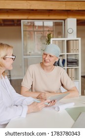 Vertical Warm-toned Portrait Of Smiling Bald Woman Listening To Female Doctor Showing Info At Digital Tablet During Consultation On Alopecia And Cancer Recovery