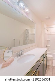 Vertical White Bathroom Interior With Round Mirror On A Stone At The Counter Of A Sleek Vanity Sink. There Is A Tub Shower With Acrylic Wall Panel And Curtain Near The Toilet