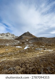 A vertical wide-angle shot of a sharp mountain peak rising above a rocky alpine meadow with scattered patches of snow under a cloudy sky at Nufenen Pass.