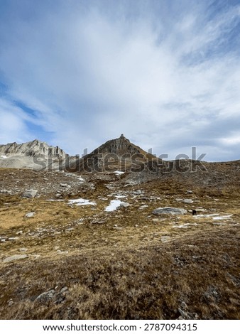A vertical wide-angle shot of a sharp mountain peak rising above a rocky alpine meadow with scattered patches of snow under a cloudy sky at Nufenen Pass.