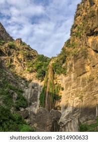 Vertical wide-angle view of a high mountain waterfall flowing through a rocky gorge under a partly cloudy sky.