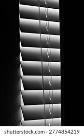 Vertical window blinds filter dim light into a dark interior, creating narrow shadows that reflect nightshift fatigue, isolation, and quiet depressive stillness.