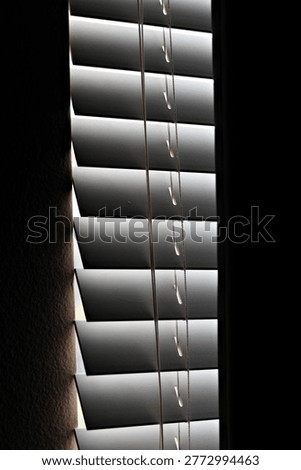 Vertical window blinds filter dim light into a dark interior, creating narrow shadows that reflect nightshift fatigue, isolation, and quiet depressive stillness.