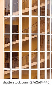 Vertical windows reflect staircase inside. White frames divide glass panes clearly. Metal railings appear within reflections. Interior stairs ascend through mirrored glass
