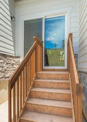 Vertical Wooden Doorsteps Of A Sliding Glass Door Of A House. House Exterior With Wooden Stairs And Railings, And An Exterior Of Gray Vinyl Wood And Stone Veneer Sidings.