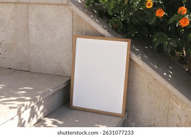 Vertical wooden picture frame leaning against stone stairway in sunlight. Textured beige tiles background, orange flowers, green leaves. Summer outdoor seeting, natural, minimal backdrop Poster mockup