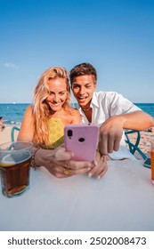 Vertical. Young couple using a smart phone to browse on internet sitting at a beach bar during their summer vacation on the coast. Boyfriend and girlfriend booking a hotel room online with a cellphone