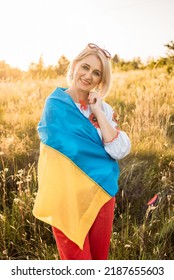 vertikal shot of happy joyfull mature woman wearing traditional Ukrainian embroidered shirt and holding ukrainian flag in countryside during beautiful sunset