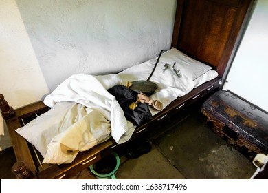 Very Antique, Messy, And Dirty Bedroom With Weathered Wooden Furnitures And White Walls