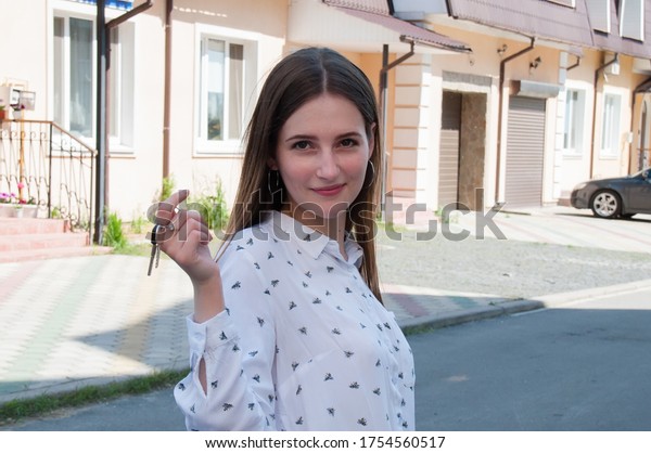 Very beautiful young woman with the keys\
to a new apartment or house in a sleeping wound of the city, among\
houses and cars in the summer on a sunny\
day