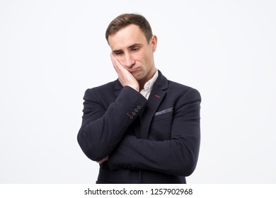 Very Bored Adult Italian Man In Suit Holding Hand On Cheek, Looking Tired And Sick, Over Gray Background.