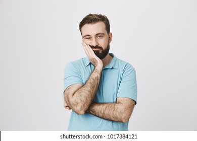 Very bored adult man with beard holding hand on cheek while support it with another crossed hand, looking tired and sick, over gray background. Father attends parents day at school