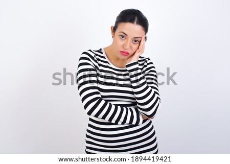 Very bored Young beautiful woman wearing stripped t-shirt against white background holding hand on cheek while support it with another crossed hand, looking tired and sick,