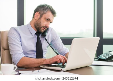 Very busy. Confident mature man in shirt and tie working at laptop and talking on the phone while sitting at his working place