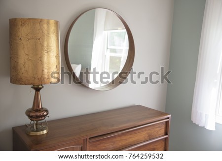 A very cool mid-century modern gold lamp sits on a teak dresser next to a circular mirror in a bedroom with grey walls