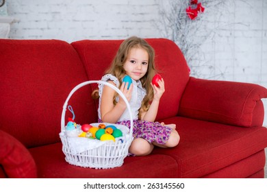 Very Cute Little Girl Sitting On A Red Couch In The Room And Holding Two Easter Egg Near Basket Full Of Colorful Eggs