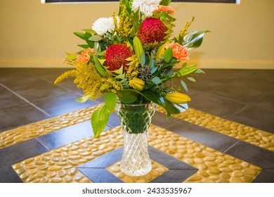 A very delightful bouquet of mixed summer flowers including red proteas,white chrysanthemums and pretty carnations is sitting in a crystal vase on a tiled floor inset with yellow pebble stones.