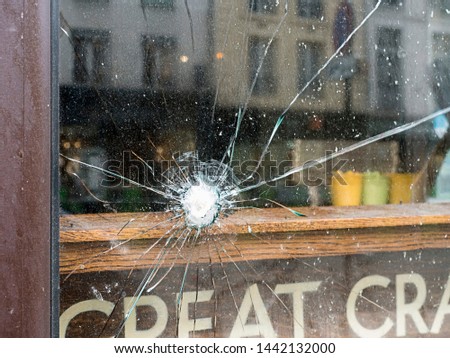The very dirty exterior window of a building shows damage from a strike of some kind, and spidery radiating cracks extending. Unidentifiable buildings across the street are blurred in the reflection.