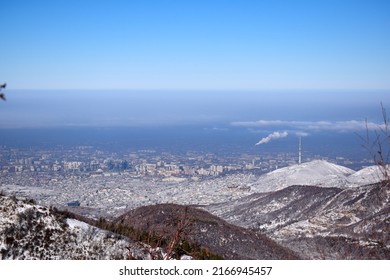 Very distant aerial view of winter city covered by thick smog, with signs of polluting smoke plumes coming into air. Aerial view of winter Almaty in morning, shot from mountains. Clear smog boundary.