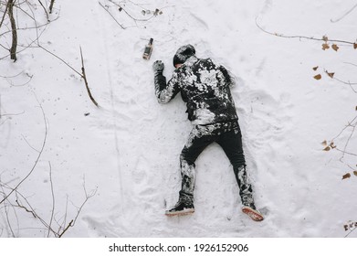 A very drunk homeless and dirty unconscious man in a black coat lies, sleeps on his stomach on white snow in a cold frosty winter with a bottle of alcohol, strong whiskey. Photography, copy space.