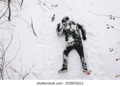 A very drunk homeless and dirty unconscious man in a black coat lies, sleeps on his stomach on white snow in a cold frosty winter with a bottle of alcohol, strong whiskey. Photography, copy space.