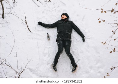 A Very Drunk Homeless Man In A Black Coat And Hat Lies, Asleep On His Back On White Snow In A Cold Frosty Winter With A Bottle Of Alcohol. Photography, Concept.