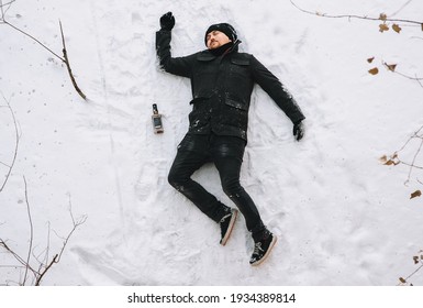 A Very Drunk Homeless Man In A Black Coat And Hat Lies, Asleep On His Back On White Snow In A Cold Frosty Winter With A Bottle Of Alcohol. Photography, Concept.