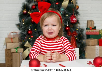 Very Emotional  And Funny Child Toddler Girl Crying Next To The Christmas Tree. Baby Girl Infant With Red Bow  Is Very Upset And Sad. Merry Christmas. Holiday Season.