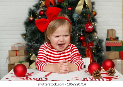 Very Emotional  And Funny Child Toddler Girl Crying Next To The Christmas Tree. Baby Girl Infant With Red Bow  Is Very Upset And Sad. Merry Christmas. Holiday Season.