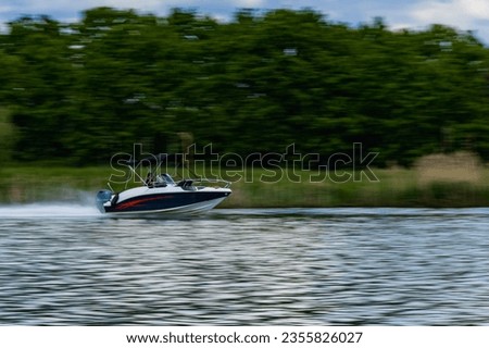 Very fast swimming small boat on wide river with green trees and bushes behind with man at the controls at cloudy morning
