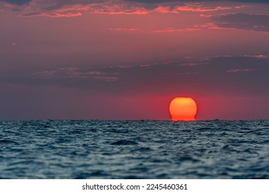 The very first moments of the sunrise, when sun rises above the sea. Sunrise over the ocean landscape with big red sun, orange sky, fluffy clouds, dark blue water. Spiritual new day natural background