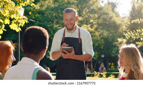 A very good looking waiter with his hair slicked back, is taking down orders in his notebook, he s talking to three women taking their orders and discussing while being in a park scenery - Powered by Shutterstock