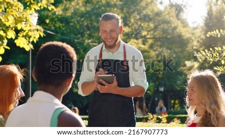 A very good looking waiter with his hair slicked back, is taking down orders in his notebook, he s talking to three women taking their orders and discussing while being in a park scenery