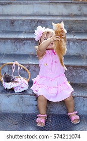 Very happy beautiful little girl in a pink dress and pink headband sitting on stairs,  holding red kitten and smiling. Causing tenderness. Vertical photo.