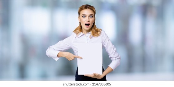 Very happy, excited surprised, astonished businesswoman with wide opened eyes, mouth, show blank paper signboard, indoors. Success in business concept. Copy space empty place for text. Astonishment.