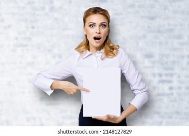 Very happy, excited surprised, astonished businesswoman with wide opened eyes, mouth, showing blank paper signboard. Success in business concept. Copy space place for text. White bricks. Astonishment.