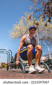 Very Happy, Healthy And Fit Young Latin Man With Weight Vest, Blue Visor And Shorts Doing Calisthenics (using Parallels) On A Street Workout Park In A Sunny Day