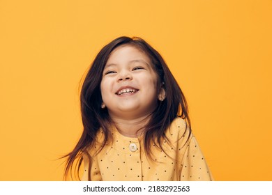 A Very Happy, Playful Girl Of Preschool Age In A Beautiful Dress Stands On An Orange Background With An Empty Space For An Advertising Layout And Opens Her Mouth Very Cheerfully From Happiness