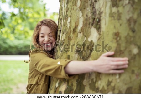 Very Happy Teen Blond Girl Hugging Huge Tree Trunk at the Park with Eyes Closed and Toothy Smile.