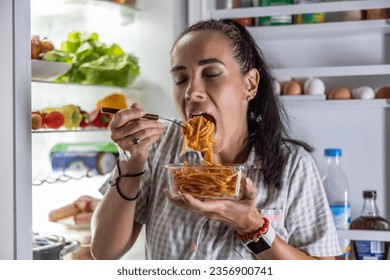 A very hungry woman in pajamas is enjoying spaghetti at the refrigerator at night.